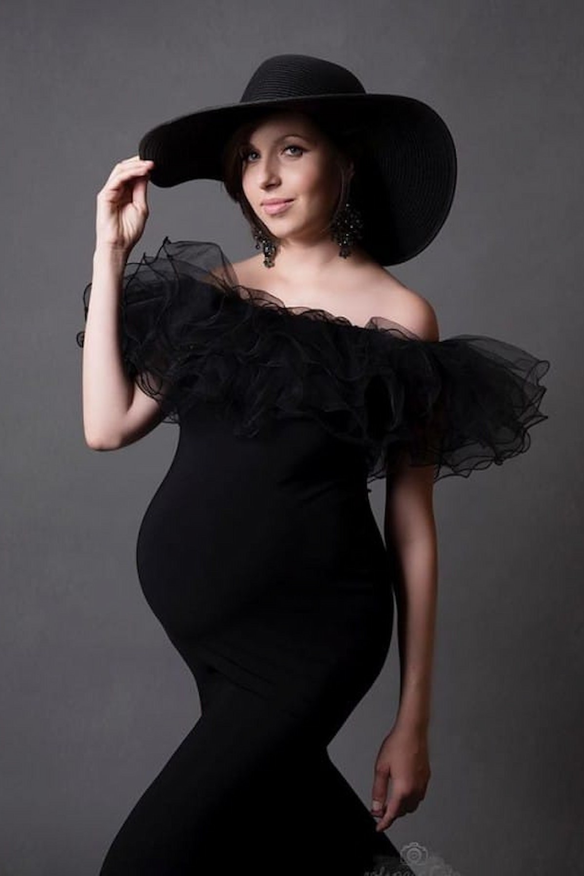 Elegant fitted maternity gown with off shoulder ruffle and wide brim hat, studio shoot