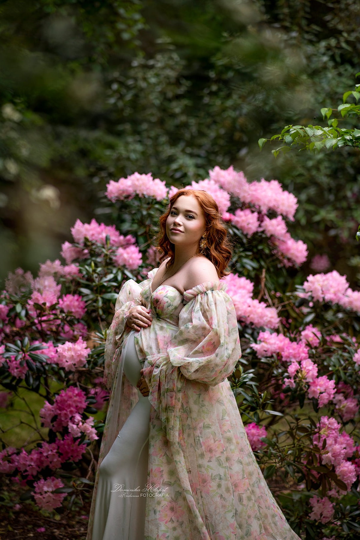 Pregnant woman in Rosa floral dress posing among pink flowers, romantic styling for maternity photoshoot.