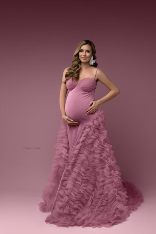 Pregnant woman wearing a dusty pink maternity gown with fitted top and voluminous ruffled tulle skirt against a studio background
